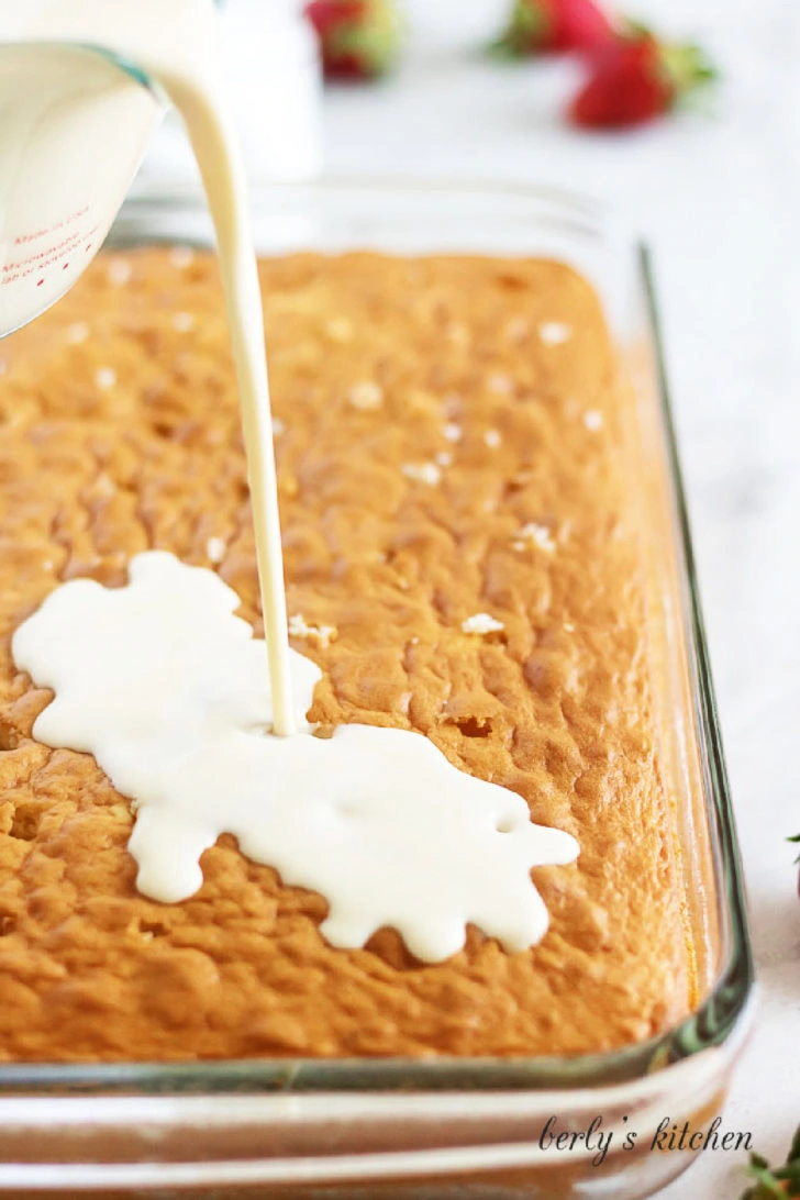 Pouring three milks mixture over baked cake in a glass baking dish.