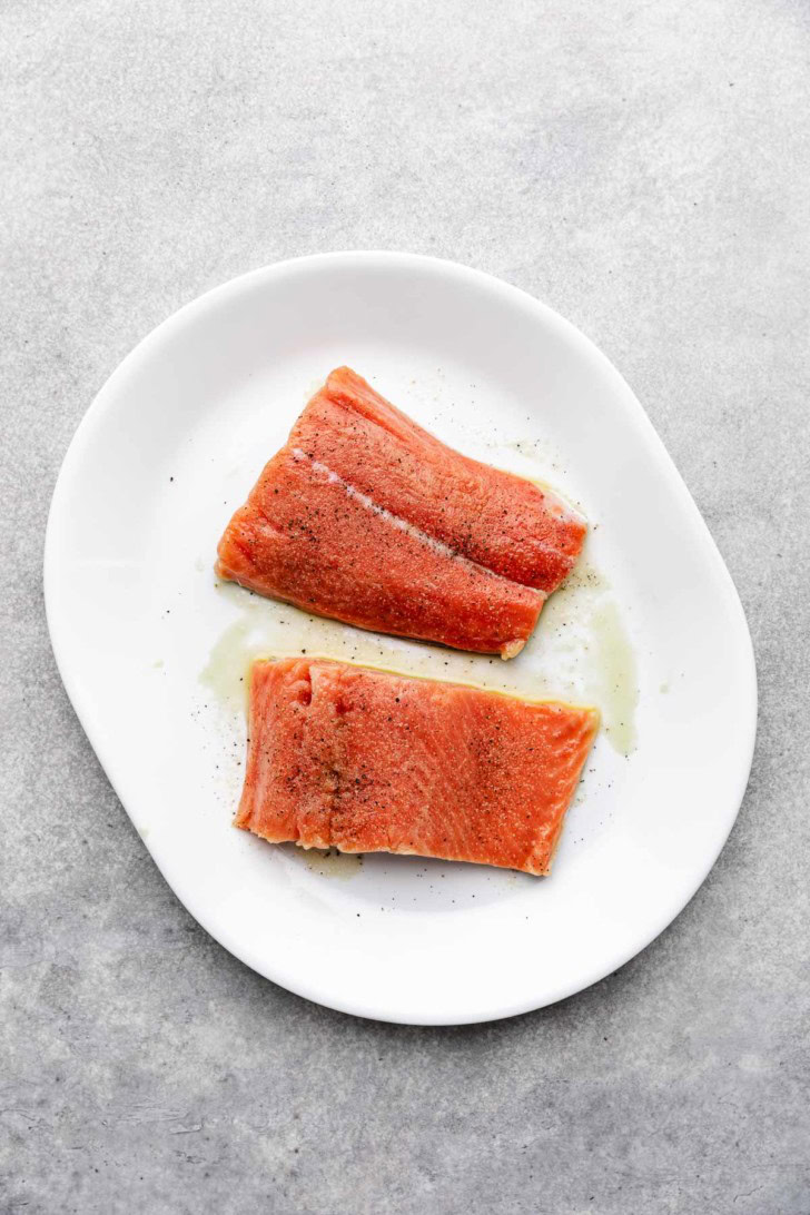 Uncooked salmon fillets with salt and pepper on a white dish.