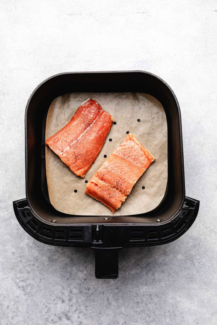 Two salmon fillets in an air fryer basket on parchment paper.