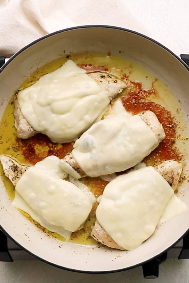 Mozzarella cheese slices melted over chicken breasts in a skillet.