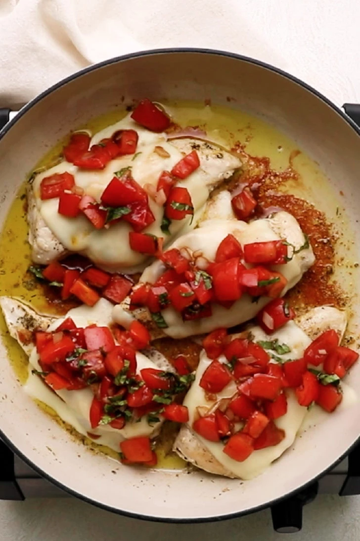 Bruschetta spooned over cheesy chicken cutlets in a pan.
