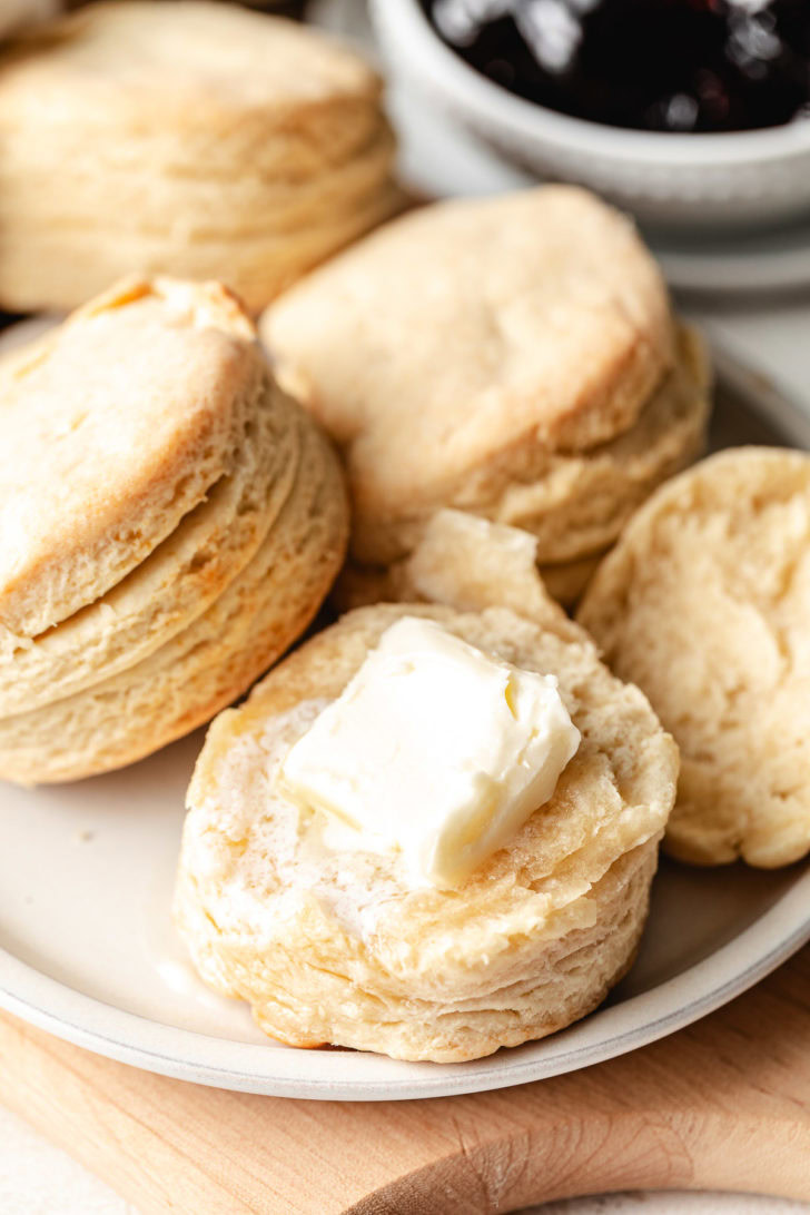 Plate of homemade buttermilk biscuits with a pat of butter on top and a bowl of jam in the background.