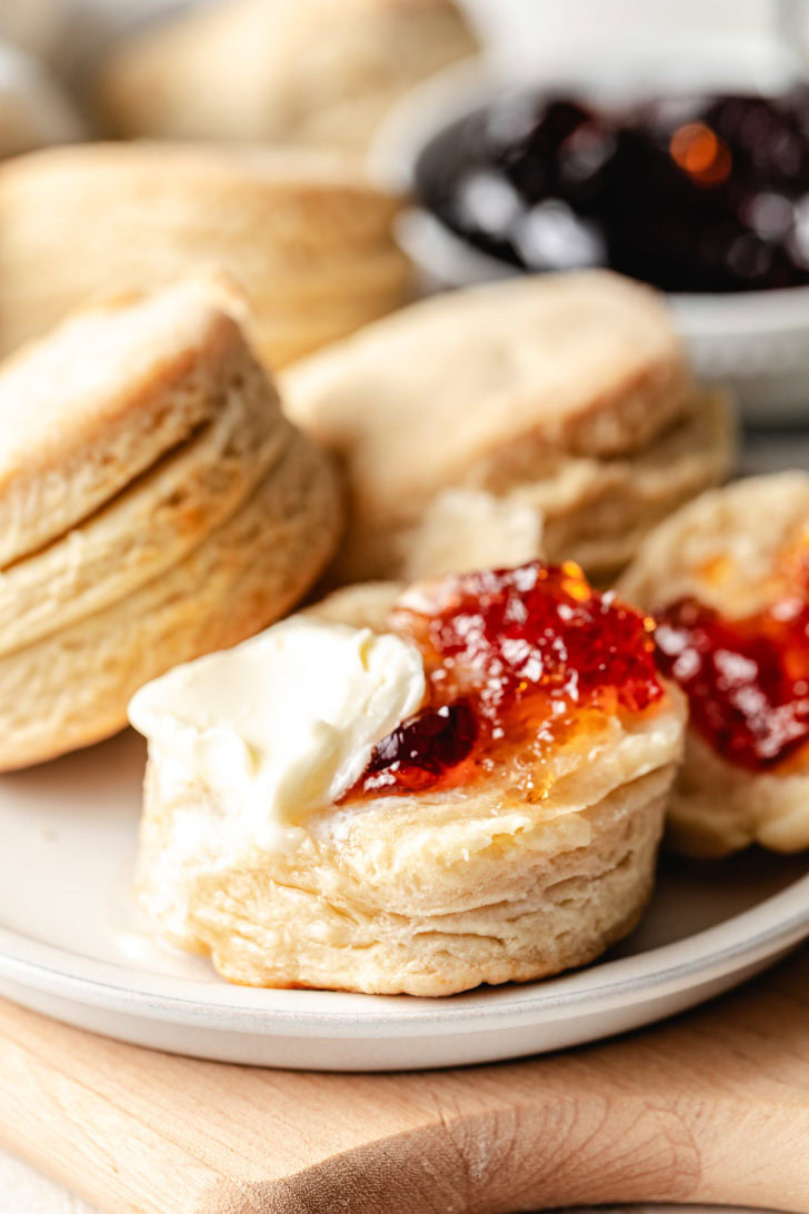 Close up of a flaky homemade biscuit with butter and strawberry jam, showing golden layers on the side.