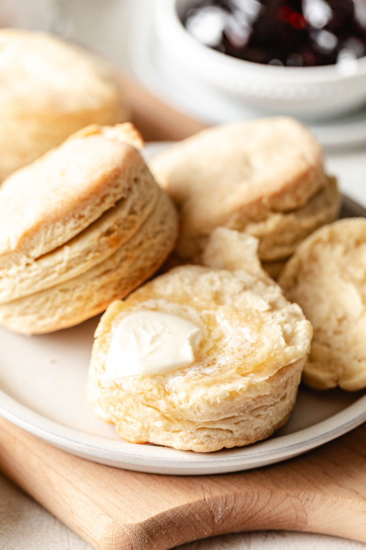 Split homemade buttermilk biscuit with a pat of butter melting inside, served on a plate with jam in the background