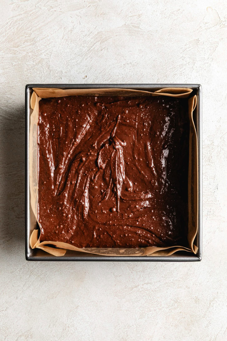 Chocolate brownie batter spread into a parchment-lined square baking pan.