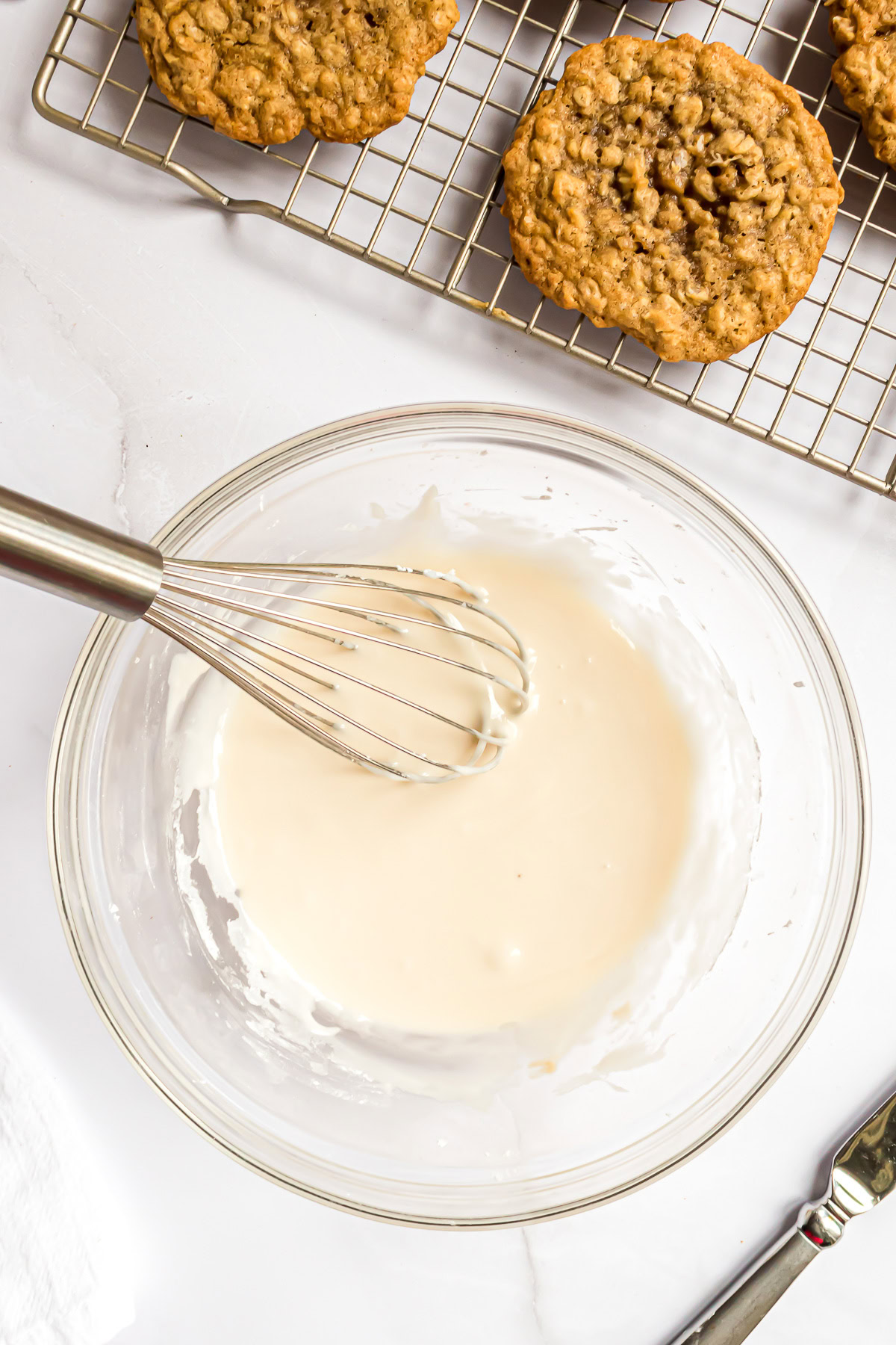 Vanilla icing mixed in a glass bowl with a whisk for iced oatmeal cookies.