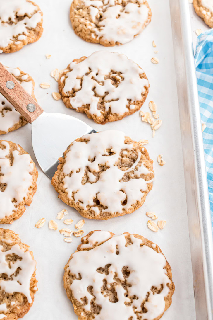 A spatula lifting an iced oatmeal cookie from a parchment-lined baking sheet.
