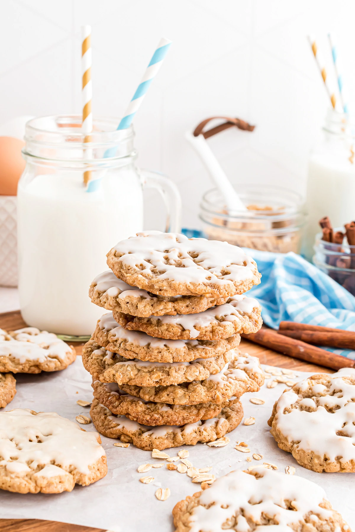 A stack of iced oatmeal cookies with jars of milk, cinnamon sticks, and oats in the background.