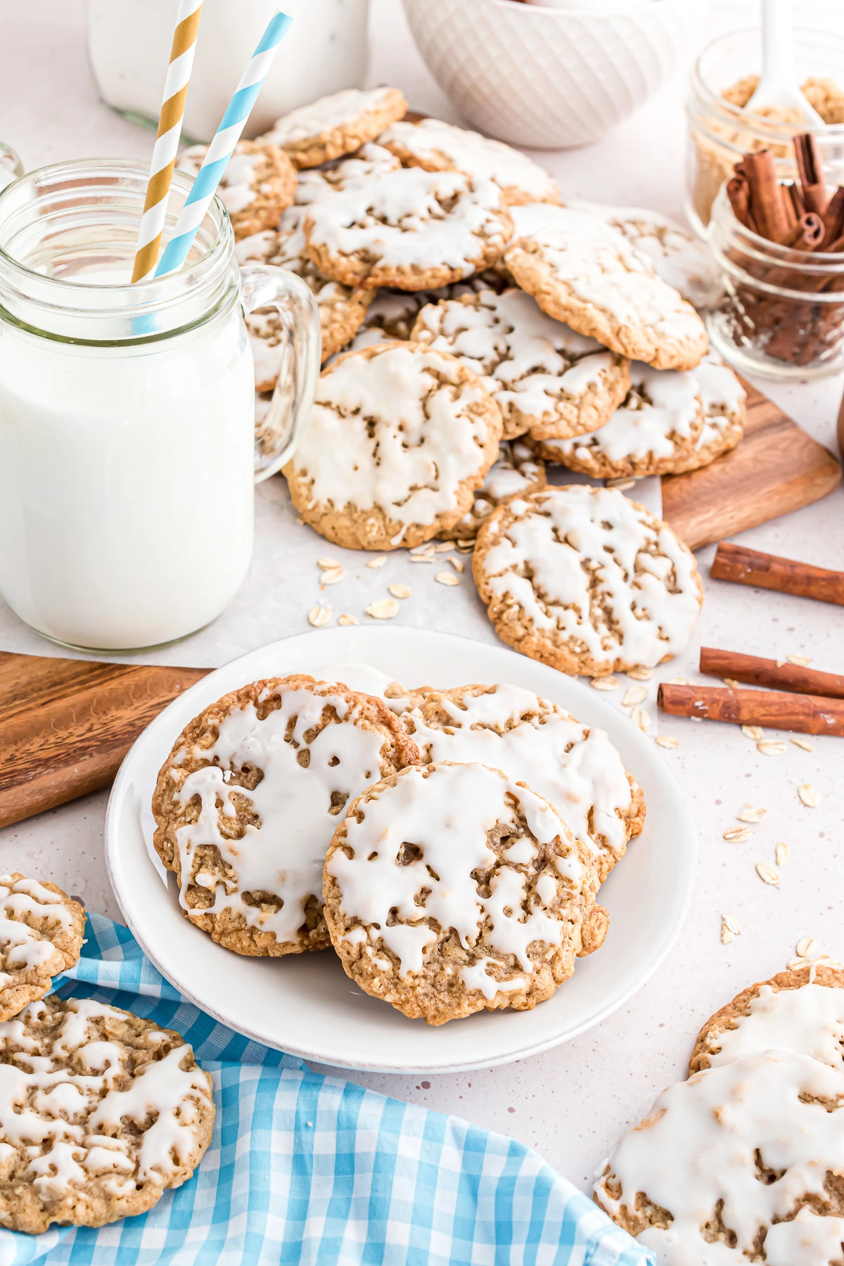 Iced oatmeal cookies on a white plate with milk, cinnamon sticks, oats, and more cookies in the background.