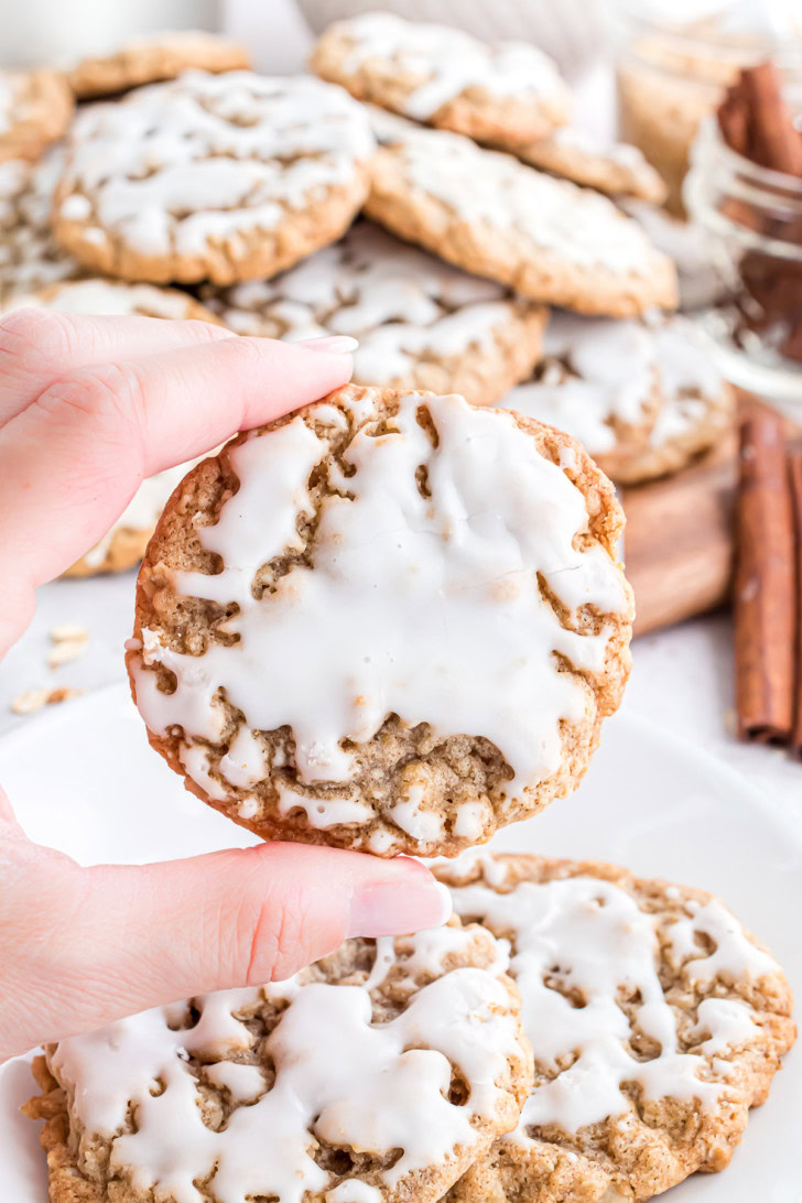 A hand holding an iced oatmeal cookie in front of more cookies and cinnamon sticks.
