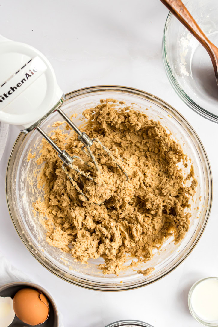 Thick oatmeal cookie dough mixed in a glass bowl with a hand mixer.