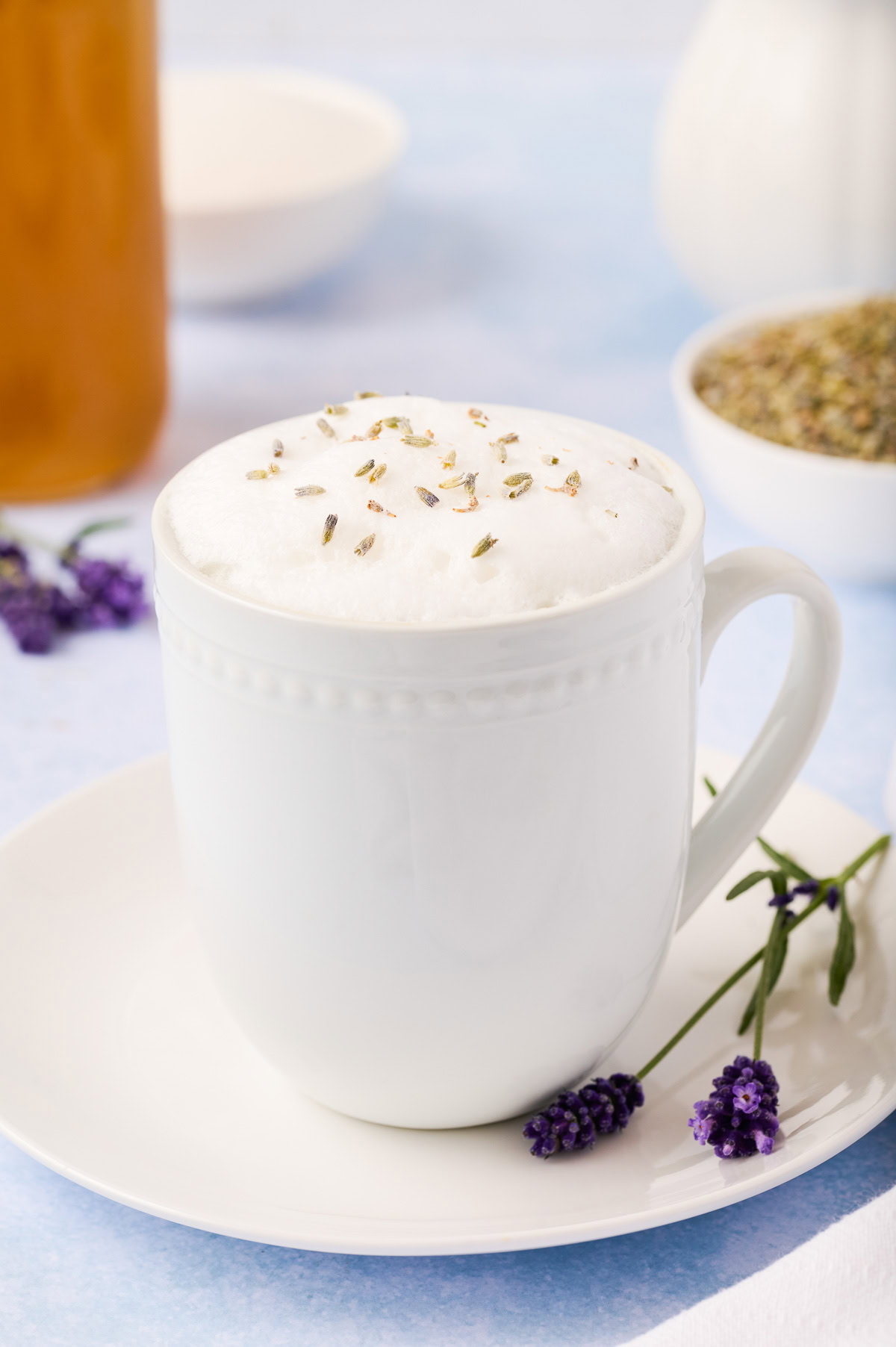 White mug of lavender latte with fresh lavender and syrup nearby.