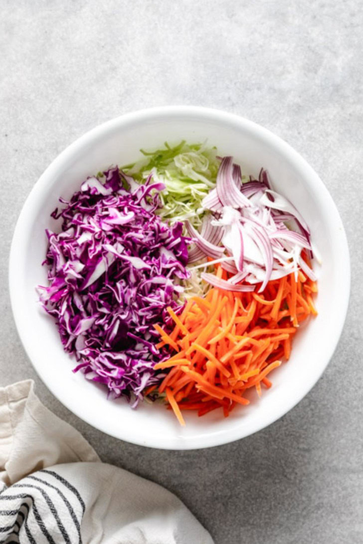 Purple and green cabbage, carrots, and onions in a white bowl.