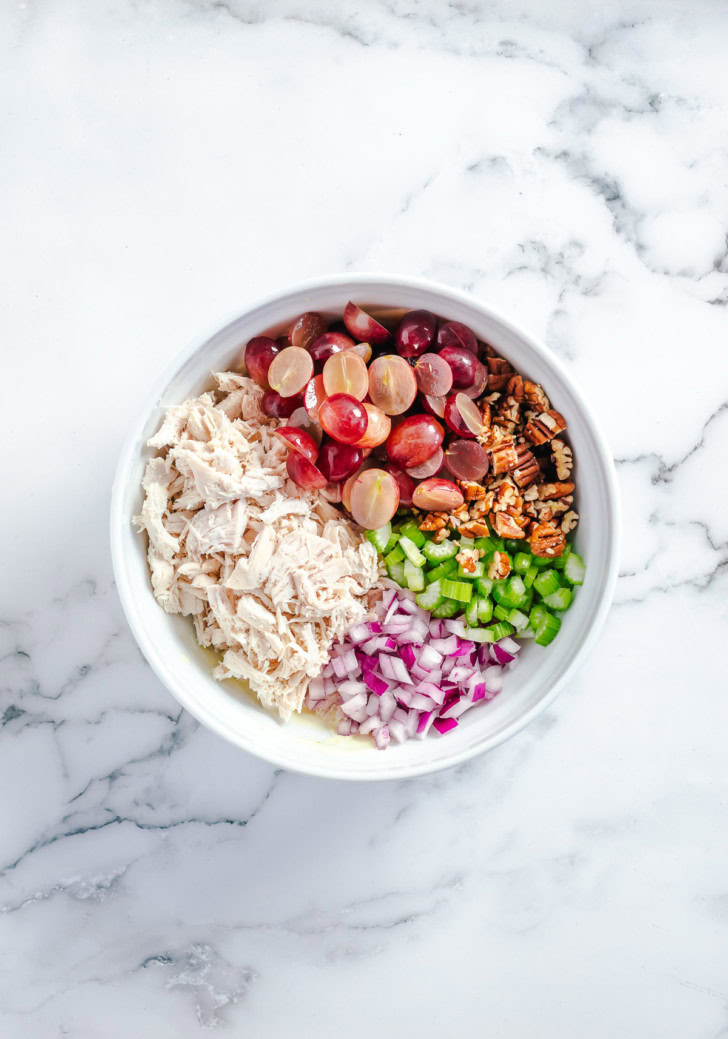 Shredded chicken, grapes, celery, pecans, and red onion arranged in a bowl.