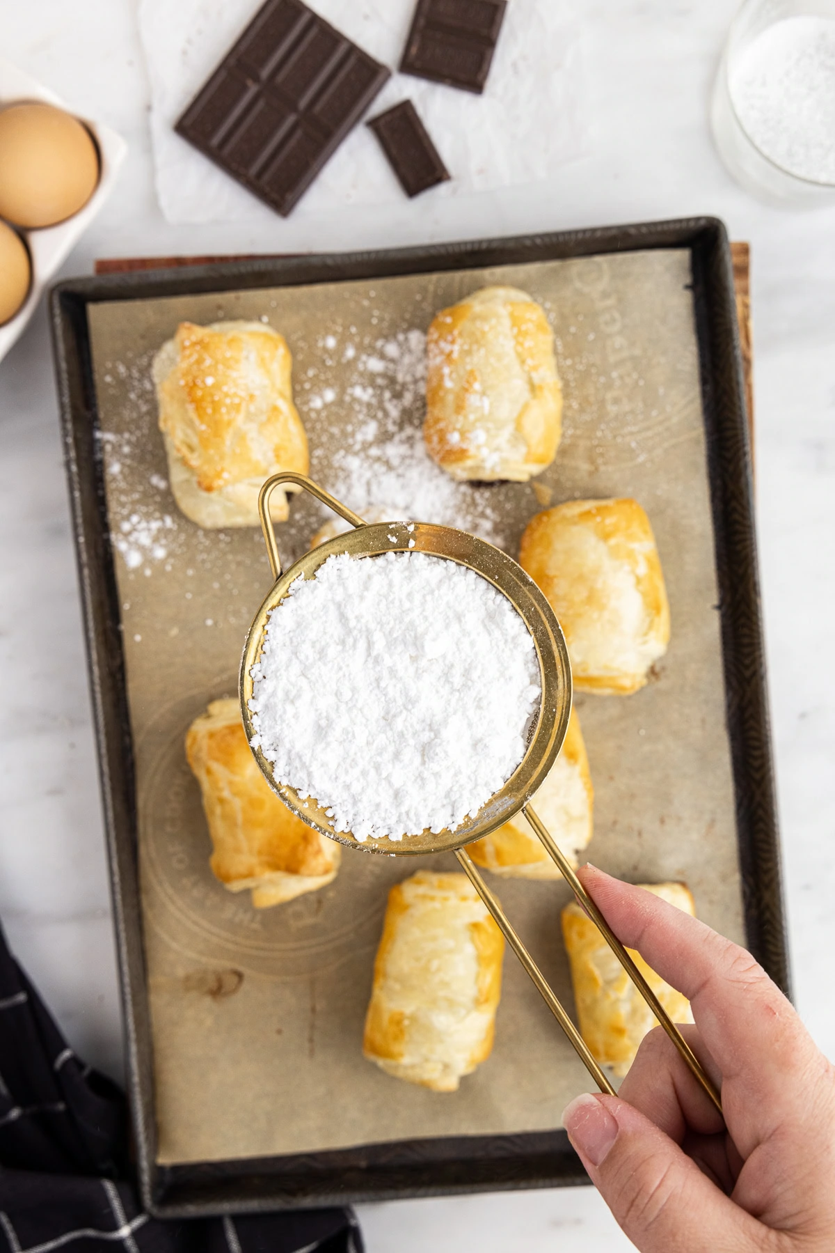 Powdered sugar being sifted over baked chocolate-filled pastries on a baking sheet.