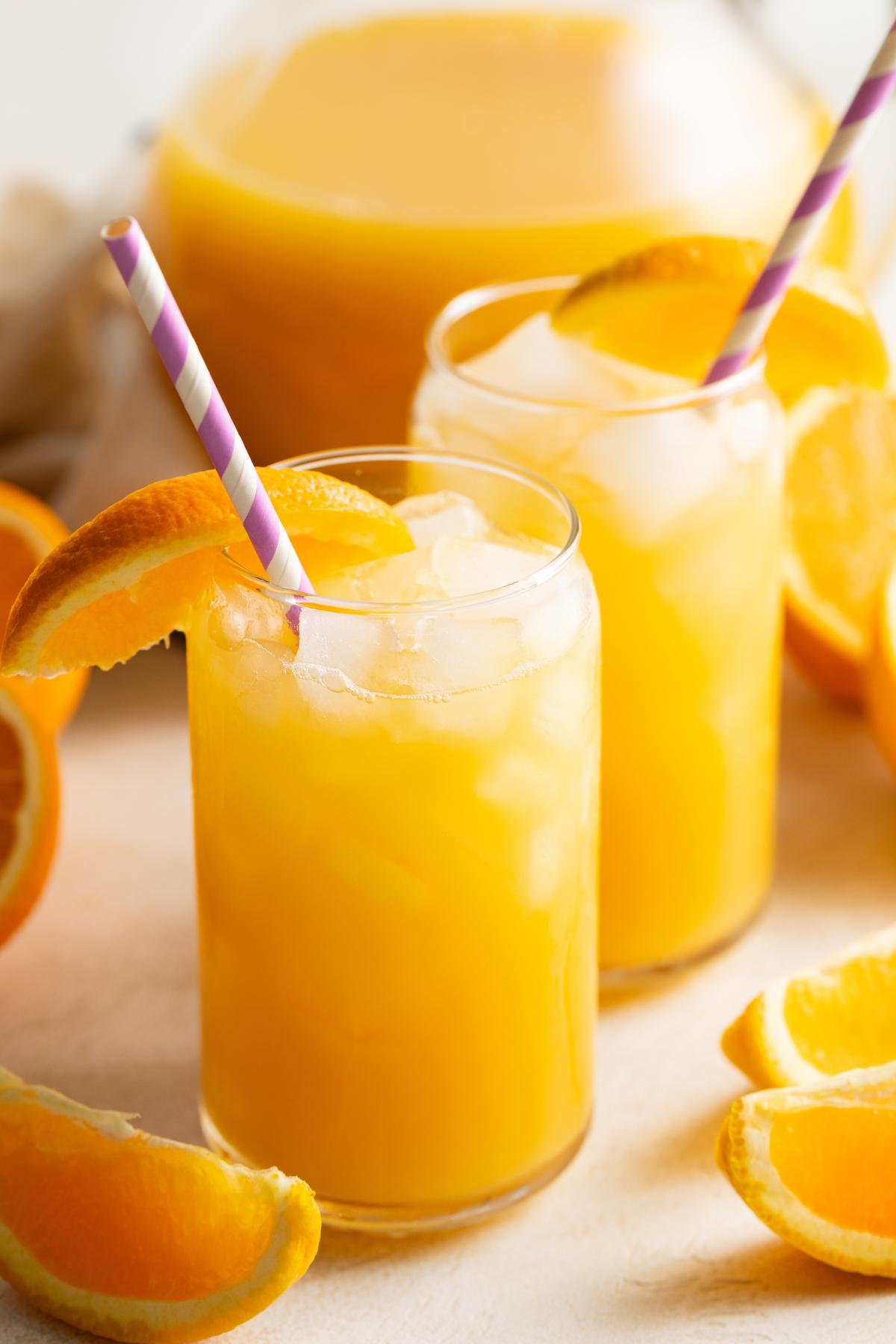 Fizzy orange juice drink served over ice with a pitcher in the background.