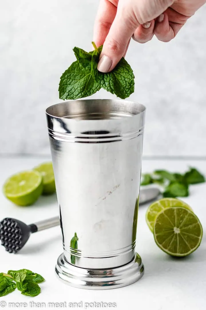 Fresh mint leaves being added to a metal cocktail shaker with limes nearby.