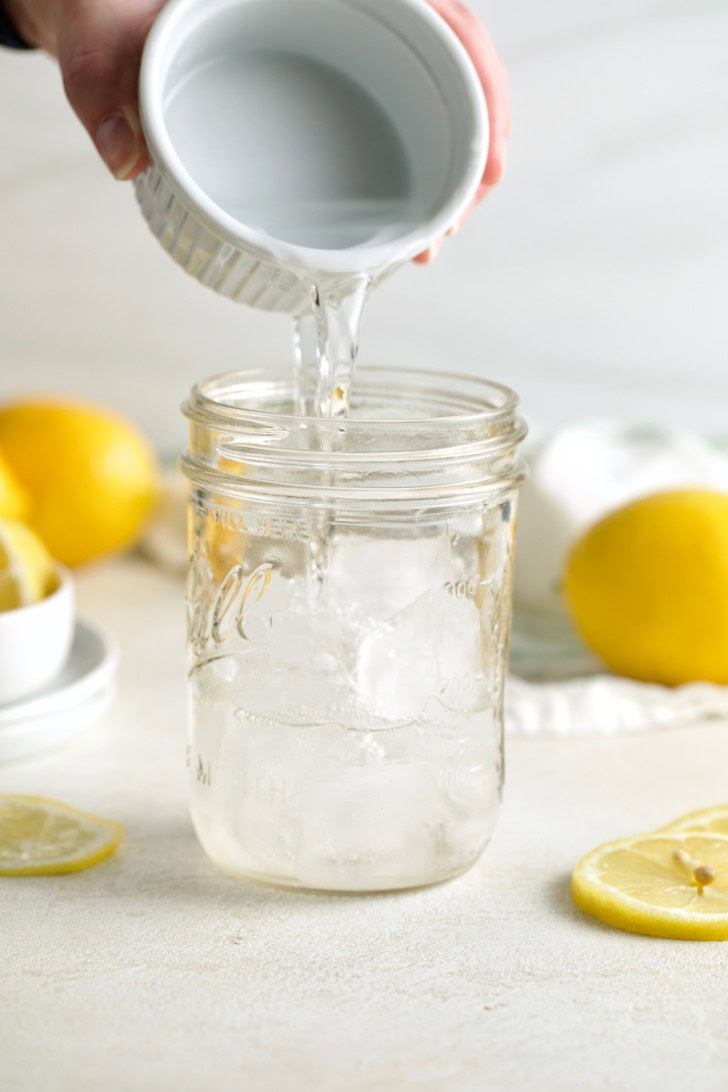 Vodka being poured into a mason jar with ice and sliced lemons nearby.
