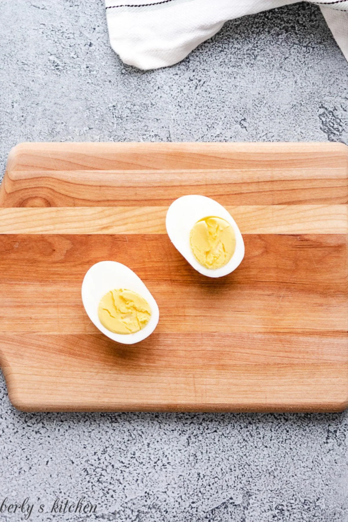 Two halved hard-boiled eggs on a wooden cutting board with a gray textured background.