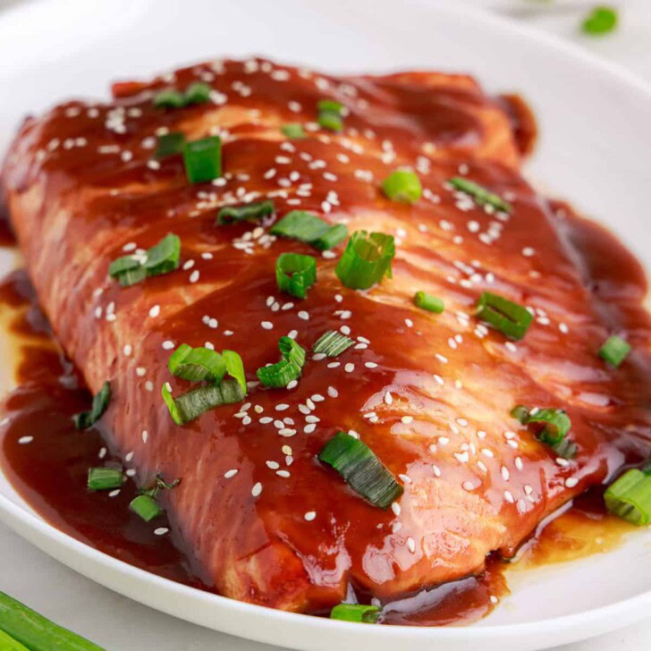 A salmon fillet glazed in teriyaki sauce and topped with sesame seeds and chopped green onions on a white plate.