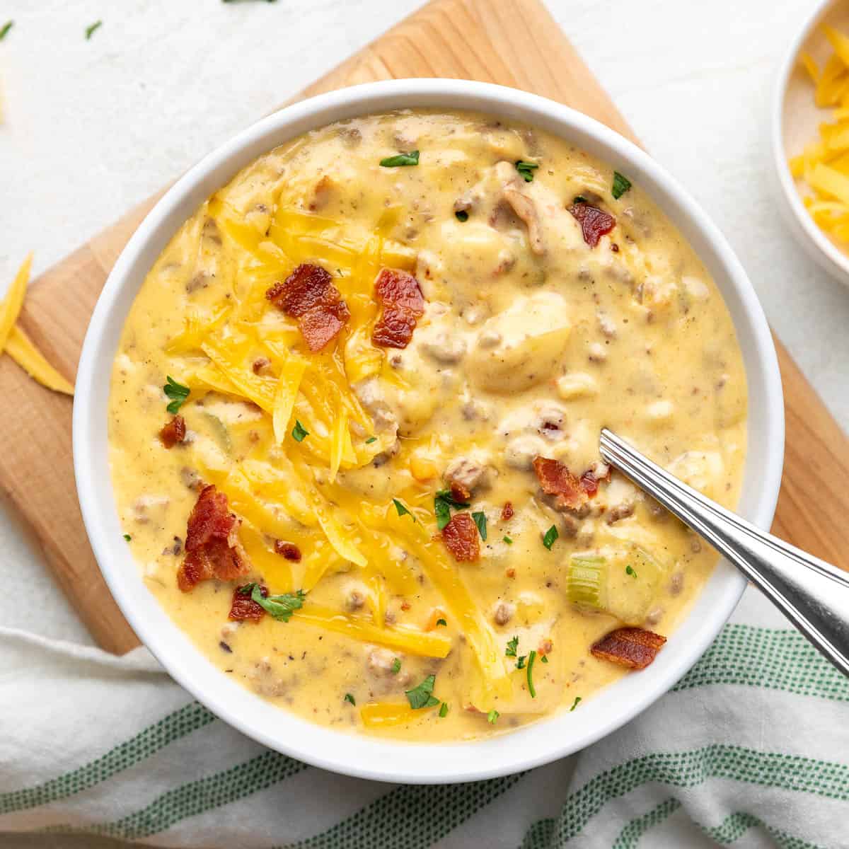 A bowl of creamy soup topped with shredded cheddar cheese, bacon bits, and parsley, with a spoon resting inside.