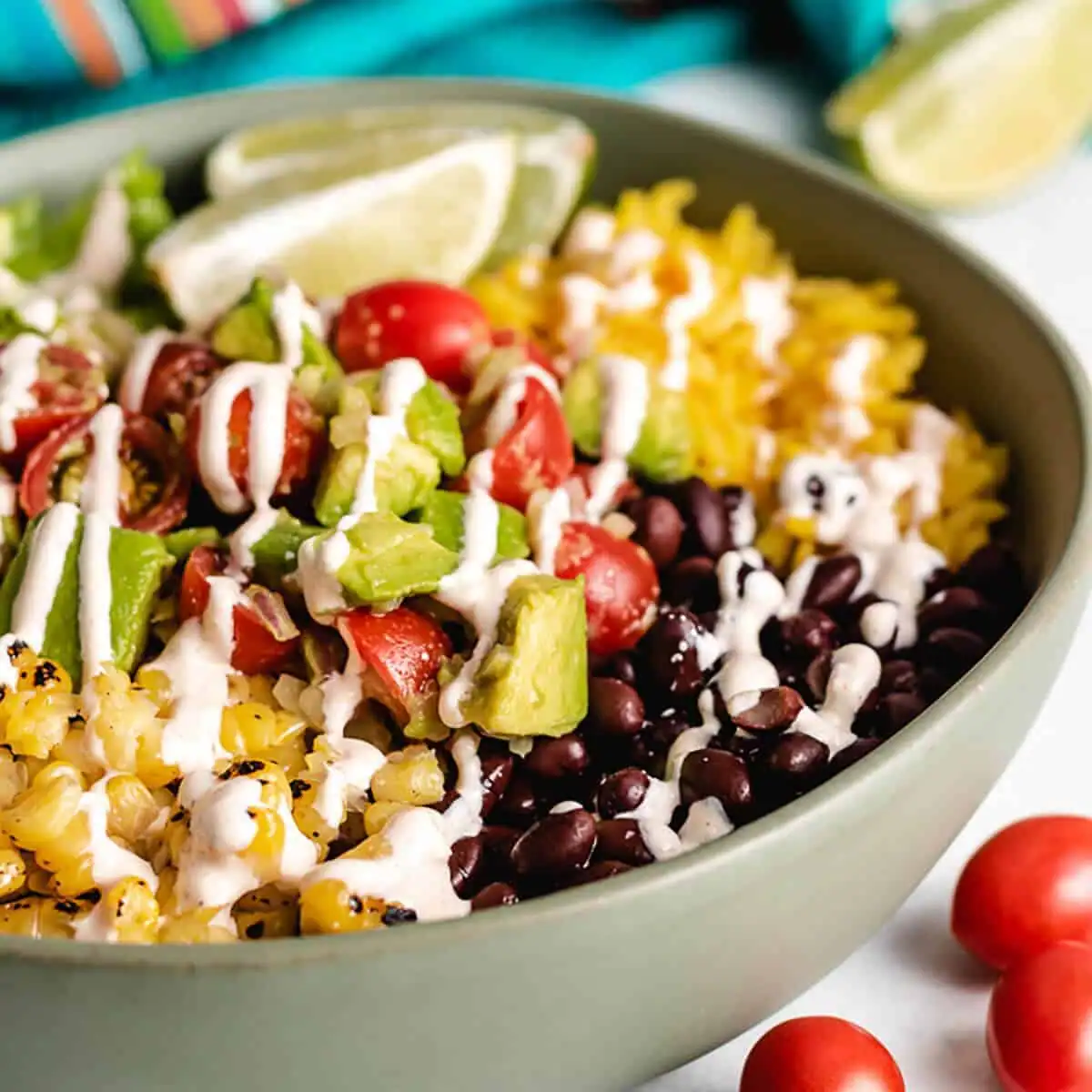 A burrito bowl in a green bowl containing black beans, seasoned rice, avocado, cherry tomatoes, and corn, drizzled with a creamy dressing and served with lime wedges.