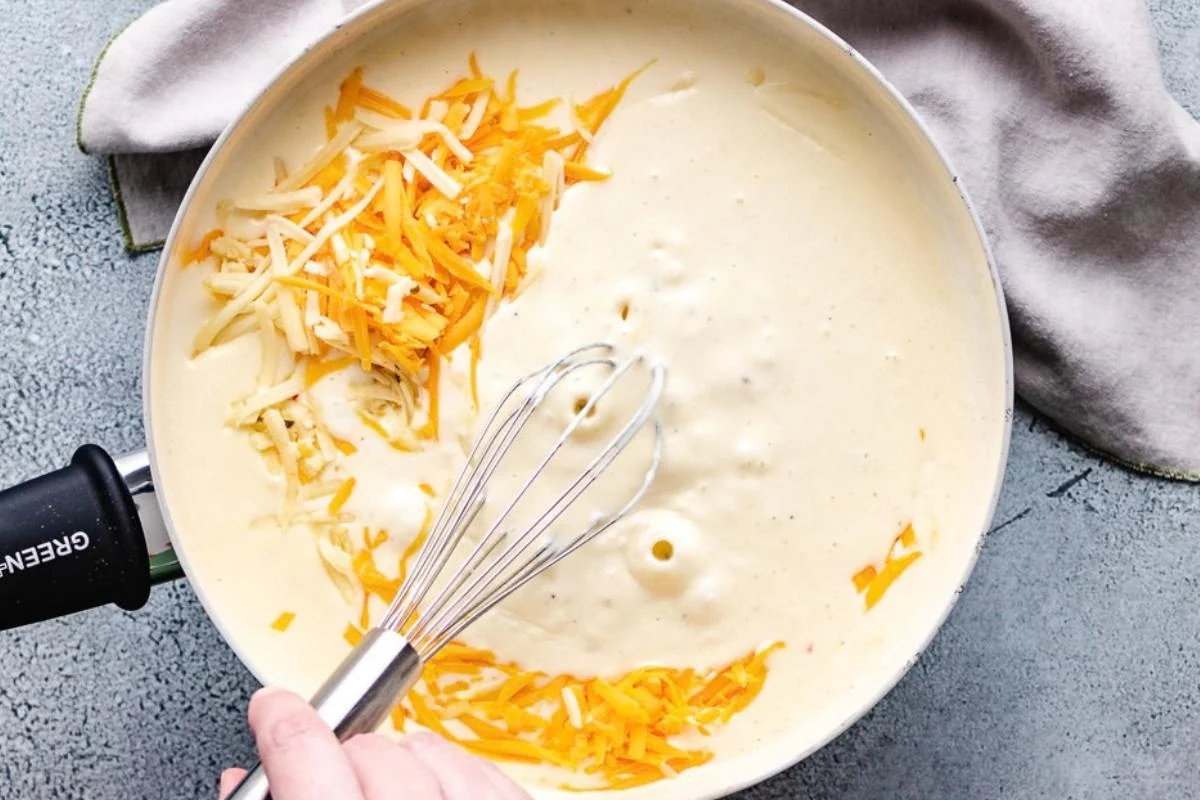 Whisk sitting in a pan of cheese sauce and shredded cheese.