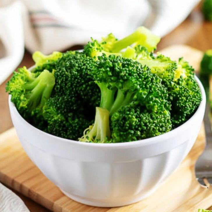 A white bowl filled with steamed broccoli florets on a wooden cutting board.