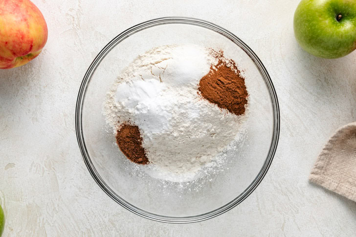 A glass bowl containing piles of flour, sugar, and cinnamon for an Irish apple cake, with a red apple and a green apple nearby.