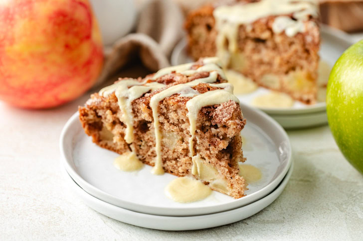 A slice of Irish apple cake drizzled with glaze on a white plate, with whole apples in the background.