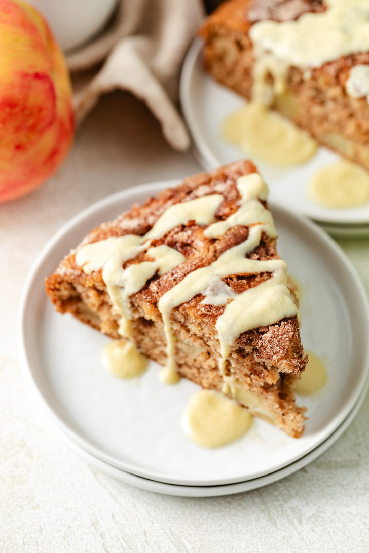 A slice of Irish apple cake drizzled with icing on a white plate, with a whole apple nearby and another slice of cake in the background.