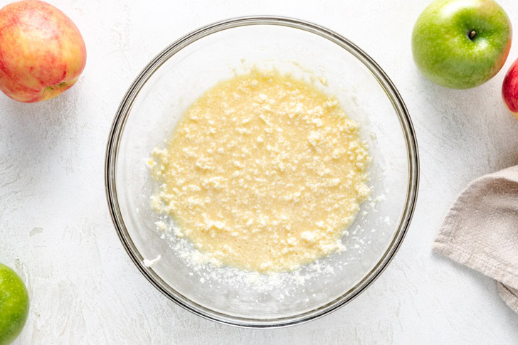 A glass bowl containing a creamed mixture of butter and sugar for an Irish apple cake, surrounded by whole apples.