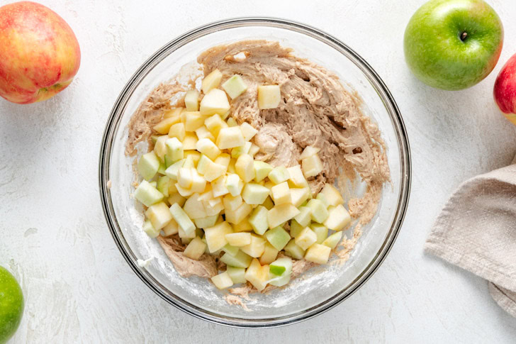 A glass bowl containing apple cake batter mixed with diced apples, surrounded by whole apples.