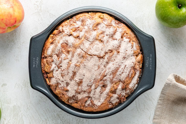 An apple cake with a cinnamon-sugar topping in a round cake pan, flanked by a red apple and a green apple.