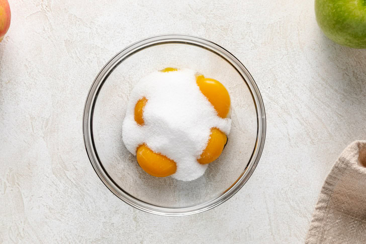 Raw egg yolks covered in white sugar in a glass mixing bowl, prepared for an Irish apple cake.