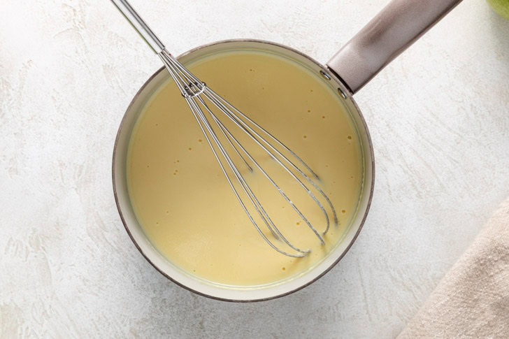 A metal whisk resting in a saucepan filled with creamy yellow custard for an Irish apple cake.